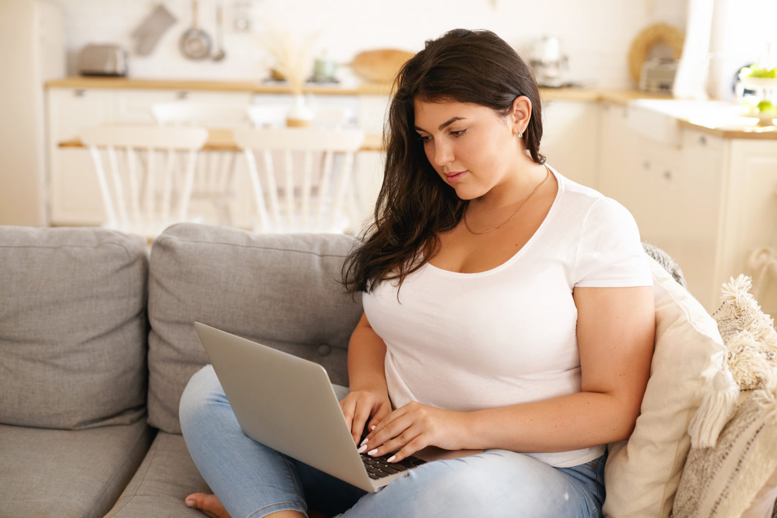 Adorable plus size student girl with loose black hair keyboarding on laptop sitting comfortably on sofa in living room, smiling, messaging friends online, using wireless high speed internet connection